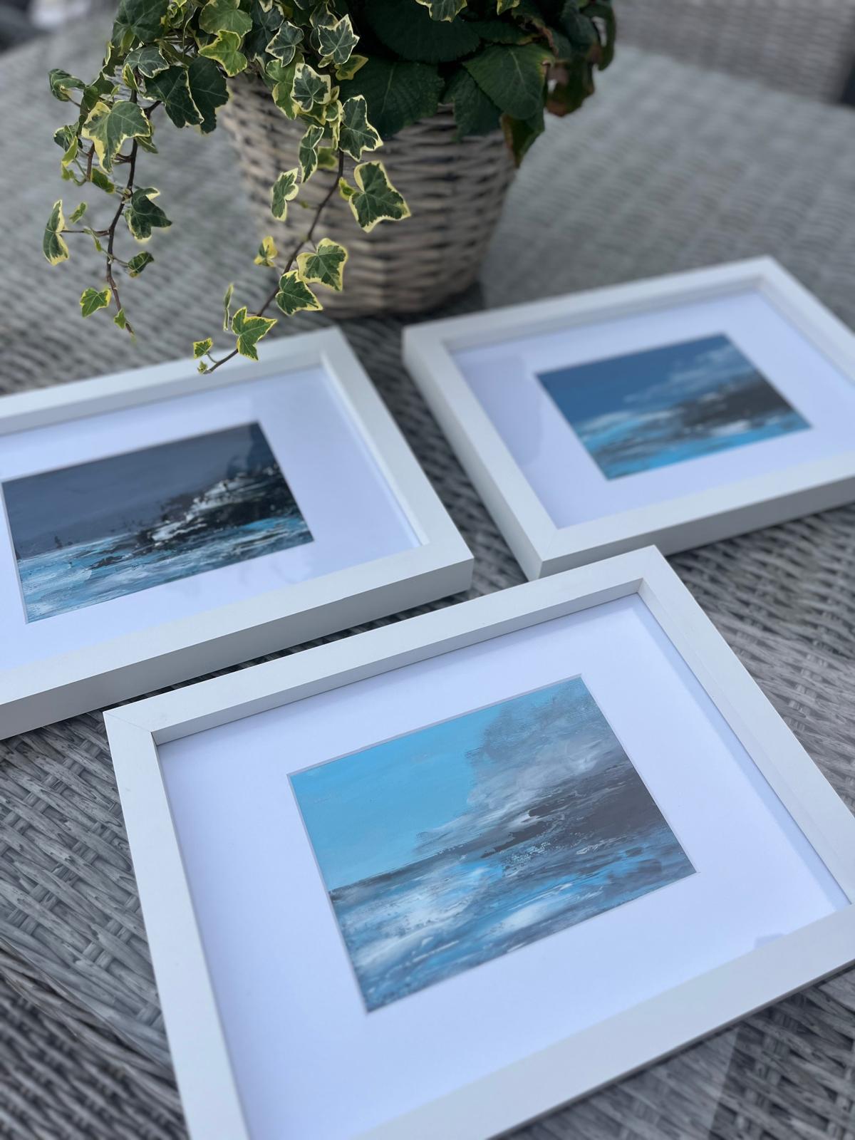 Three framed coastal artworks displayed on a woven table